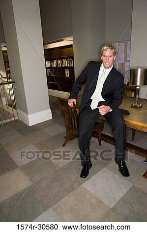 Stock Image - Portrait of a cheerful businessman sitting on table in an office. Fotosearch
