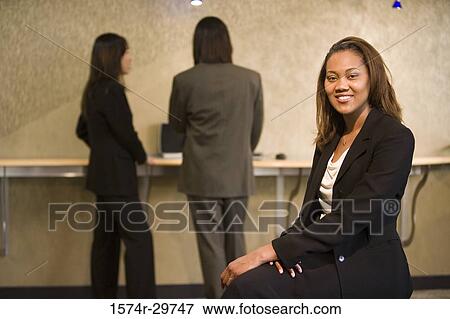 Stock Photo - Portrait of a cheerful businesswoman sitting while colleagues standing in the background. Fotosearch