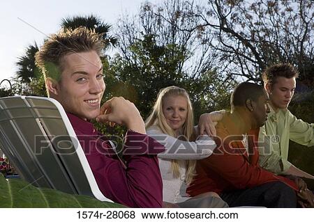 Stock Photo - Portrait of a cheerful man with hand on chin while others sitting on armchairs in background. Fotosearch