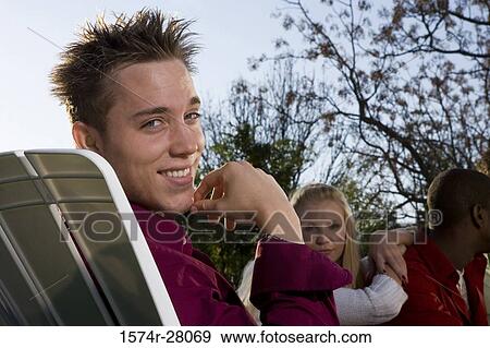 Stock Photo - Portrait of a cheerful man with hand on chin while others sitting on armchairs in background. Fotosearch