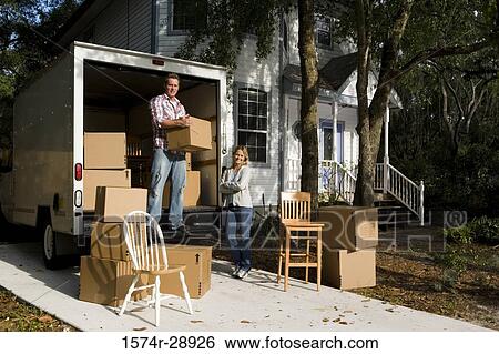 Stock Photograph - Portrait of a cheerful mid adult woman standing while mid adult man unloading chairs from the moving van. Fotosearch