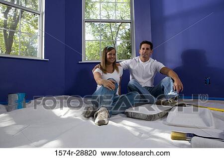 Stock Image - Portrait of a cheerful young couple sitting with paint tray and paintbrush on drop cloth in house. Fotosearch
