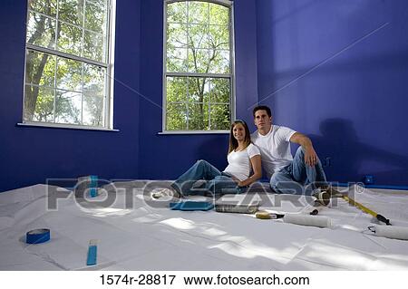Stock Photo - Portrait of a cheerful young couple sitting with paint tray and paint roller on drop cloth in house. Fotosearch