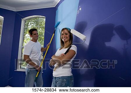 Portrait of a cheerful young woman holding paint roller with arms crossed while young man painting wall View Large Photo Image Picture - Portrait of a cheerful young woman holding paint roller with arms crossed while young man painting wall. Fotosearch