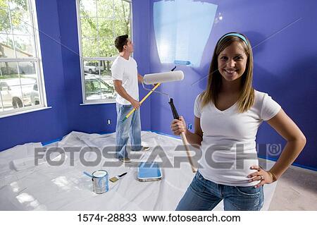 Stock Image - Portrait of a cheerful young woman holding paint roller while young man painting wall. Fotosearch