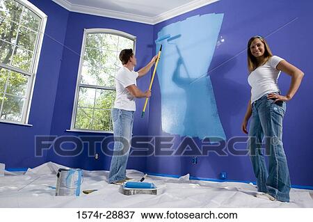 Portrait of a cheerful young woman standing with hand on hip while young man painting wall View Large Photo Image Stock Photo - Portrait of a cheerful young woman standing with hand on hip while young man painting wall. Fotosearch