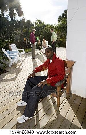 Stock Photo - Portrait of a contemplative man sitting on armchair while friends discussing in background. Fotosearch