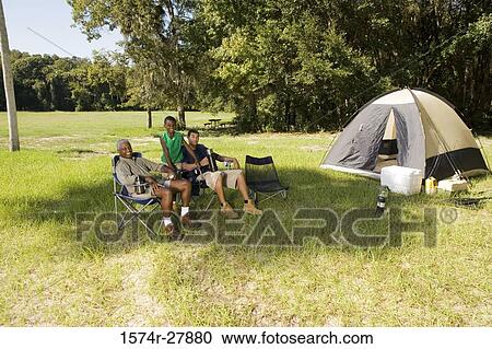 Stock Image - Portrait of a man relaxing with his friend and son on chair by tent at forest. Fotosearch