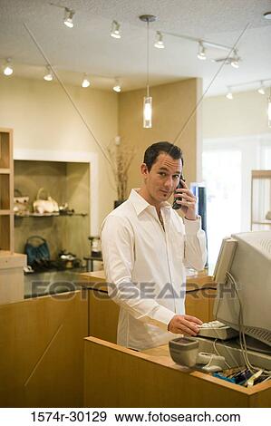 Stock Photo - Portrait of a man standing in front of a computer in a store while using phone. Fotosearch