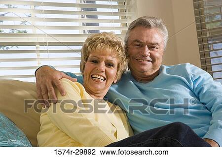 Stock Image - Portrait of a smiling mature couple sitting on a couch in living room. Fotosearch