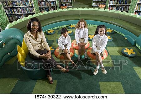 Stock Image - Portrait of a smiling teacher and schoolgirls sitting on stools in the library. Fotosearch