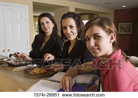 Stock Image - Portrait of a teenage girl studying while her mother and sister having food by kitchen counter. Fotosearch