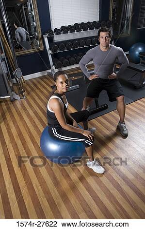 Stock Image - Portrait of a trainer sitting with a young woman with fitness ball in the gym. Fotosearch