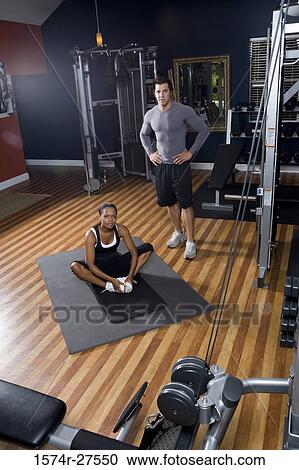 Stock Image - Portrait of a trainer standing by a young woman in the gym. Fotosearch