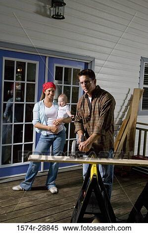 Stock Photography - Portrait of a young man cutting plank with circular saw while young woman holding baby. Fotosearch