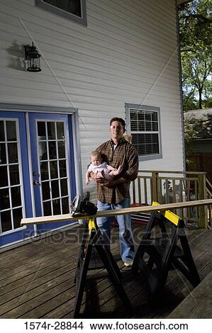 Picture - Portrait of a young man with baby standing on wooden plank by house. Fotosearch