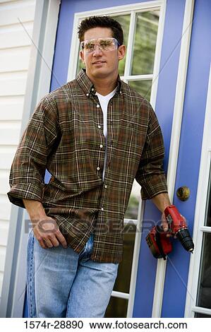 Stock Image - Portrait of a young man with drill standing by door. Fotosearch