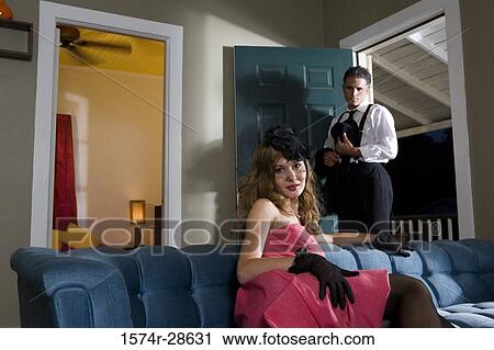 Stock Image - Portrait of a young prostitute in a red dress sitting on sofa while mid adult man standing by door. Fotosearch