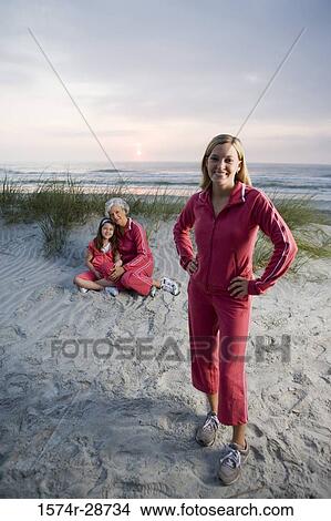 Picture - Portrait of a young woman standing while mature woman sitting with her granddaughter at the beach. Fotosearch