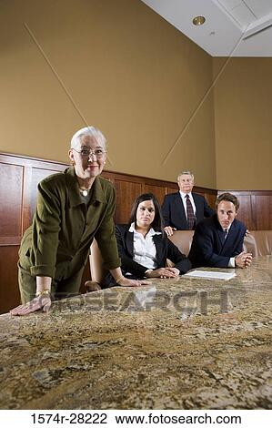Stock Image - Portrait of businesswoman leaning on conference table while colleagues sitting on chairs in background. Fotosearch