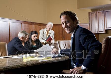 Portrait of cheerful businessman sitting by conference table while colleagues discussing in the background View Large Photo Image Stock Photography - Portrait of cheerful businessman sitting by conference table while colleagues discussing in the background. Fotosearch