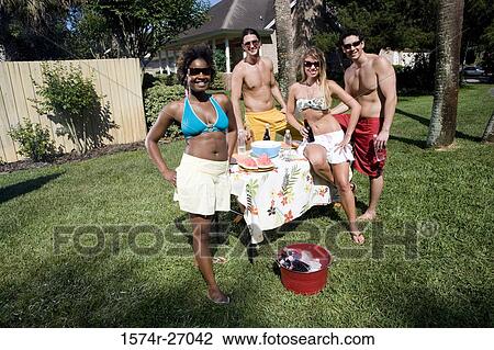 Stock Image - Portrait of cheerful friends standing by table on lawn. Fotosearch