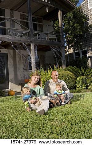 Stock Photo - Portrait of cheerful parents sitting with their children in front of a house. Fotosearch