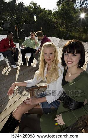 Stock Photo - Portrait of cheerful women sitting on armchairs while men discussing in background. Fotosearch