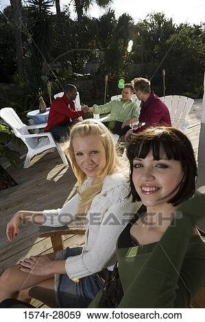 Portrait of cheerful women sitting on armchairs while men discussing in background View Large Photo Image Stock Photo - Portrait of cheerful women sitting on armchairs while men discussing in background. Fotosearch