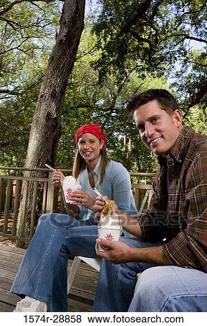 Stock Photo - Portrait of cheerful young couple sitting together on armchairs and eating. Fotosearch