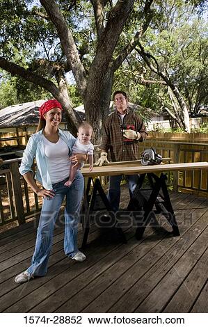Stock Image - Portrait of cheerful young woman carrying baby while young man standing with drill. Fotosearch