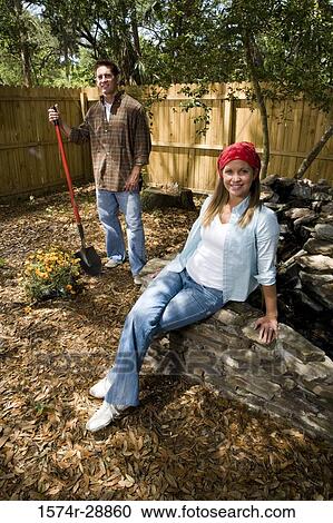 Stock Image - Portrait of cheerful young woman smiling while young man holding shovel. Fotosearch