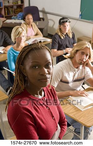 Stock Image - Portrait of female teacher with arms crossed while students reading book. Fotosearch
