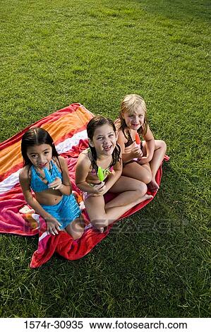Stock Photography - Portrait of girls sitting on a towel and eating flavored ice. Fotosearch