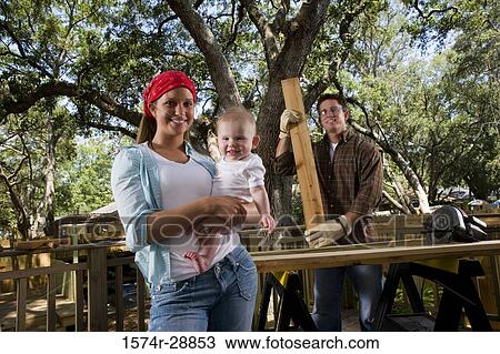 Stock Image - Portrait of young woman carrying baby while young man holding wood plank. Fotosearch