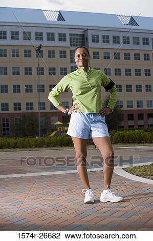 Stock Image - Portrait of young woman in sports clothing standing on walkway with building in the background. Fotosearch