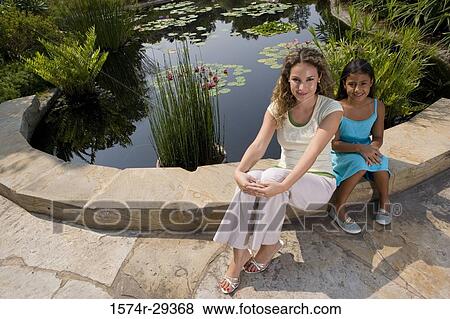 Stock Photo - Portrait of young woman with her daughter sitting by pond. Fotosearch