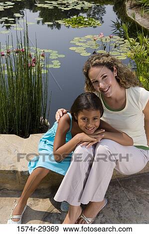 Stock Photo - Portrait of young woman with her daughter sitting by pond. Fotosearch