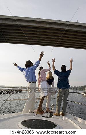 Stock Photograph - Rear view of friends standing at bow of a boat with arms raised and waving at bridge. Fotosearch