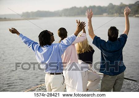 Stock Image - Rear view of friends standing with arms raised on deck of a boat. Fotosearch