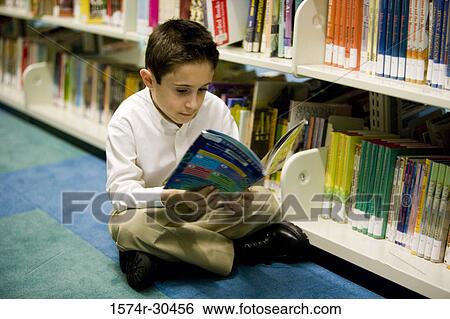 Stock Photograph - Schoolboy sitting on carpet and reading a book in the library. Fotosearch