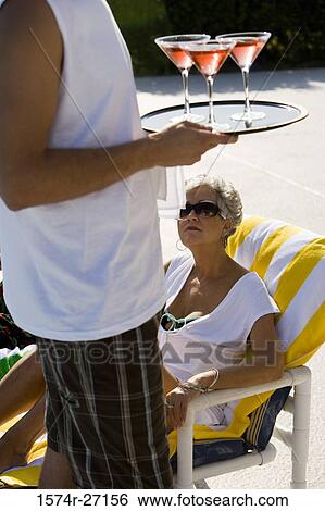 Stock Photograph - Senior woman sitting on reclining chair while man holding martini glasses. Fotosearch