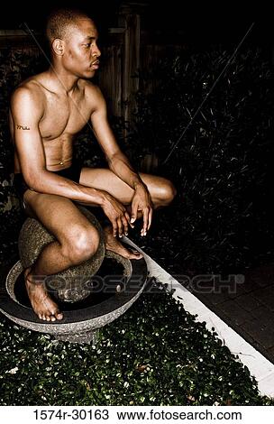 Stock Image - Shirtless young man sitting on a round structure and looking away. Fotosearch