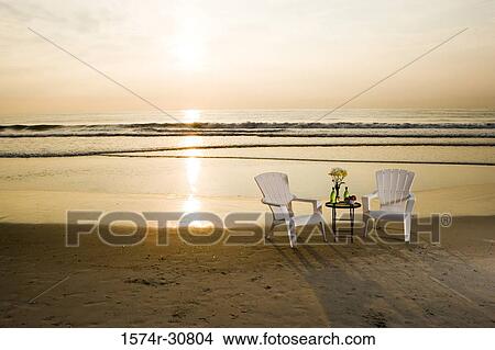 Table with chairs arranged on the beach with sunset in background View Large Photo Image Picture - Table with chairs arranged on the beach with sunset in background. Fotosearch