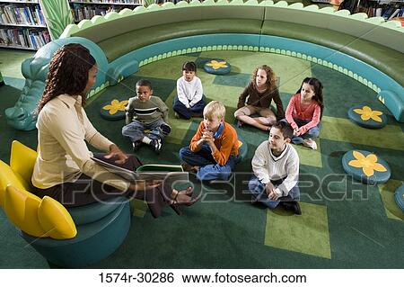 Stock Photograph - Teacher showing book to students sitting on floor in the library. Fotosearch