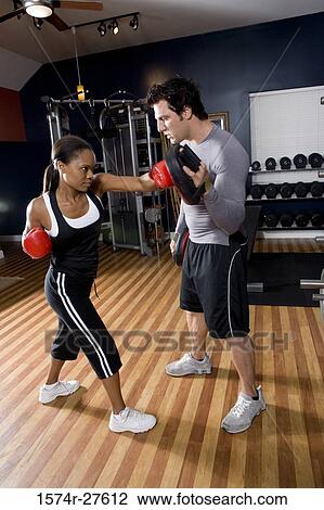 Stock Image - Trainer instructing a young woman with boxing equipment in the gym. Fotosearch