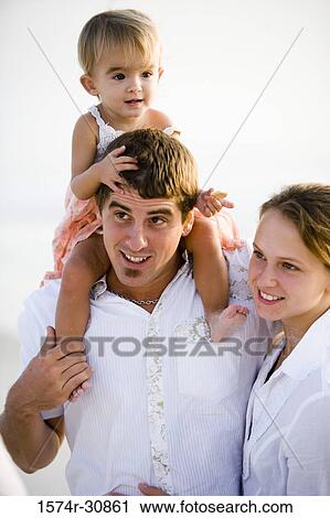 Stock Image - Young couple with girl sitting on father's shoulder at beach. Fotosearch