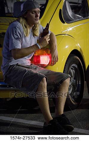 Young man looking at a bottle by a car at the parking area View Large Photo Image Stock Image - Young man looking at a bottle by a car at the parking area. Fotosearch