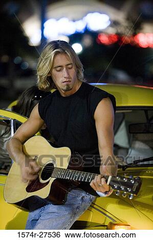 Stock Photograph - Young man playing guitar by a car at the parking area. Fotosearch