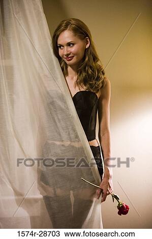 Stock Image - Young prostitute in pantyhose holding a rose while standing by curtain. Fotosearch
